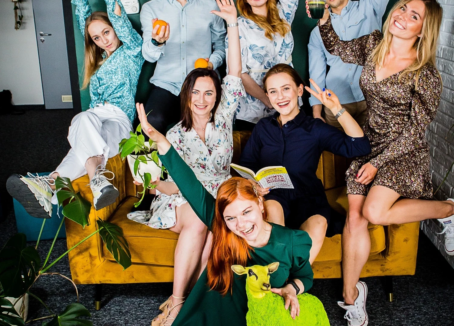 Grupa radosnych osób pozuje w kreatywnym biurowym otoczeniu przed podświetlonym logo 'Harbingers'. Wszyscy są uśmiechnięci, unoszą ręce w geście entuzjazmu, trzymają różne przedmioty, jak książka, kubek czy pomarańcze. Na pierwszym planie kobieta w zielonej sukience siedzi na podłodze, obejmując neonowo-zieloną figurkę jelenia. Zdjęcie symbolizuje pozytywną energię, kreatywność i zgraną atmosferę zespołu.