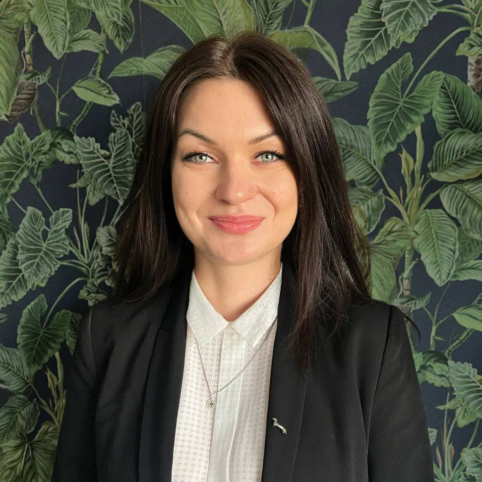 Portret kobiety w czarnym żakiecie i białej bluzce, na tle ściany z motywem roślinnym. Specjalistka ds. content marketingu, opracowująca strategie oparte na danych i analizie ruchu organicznego.