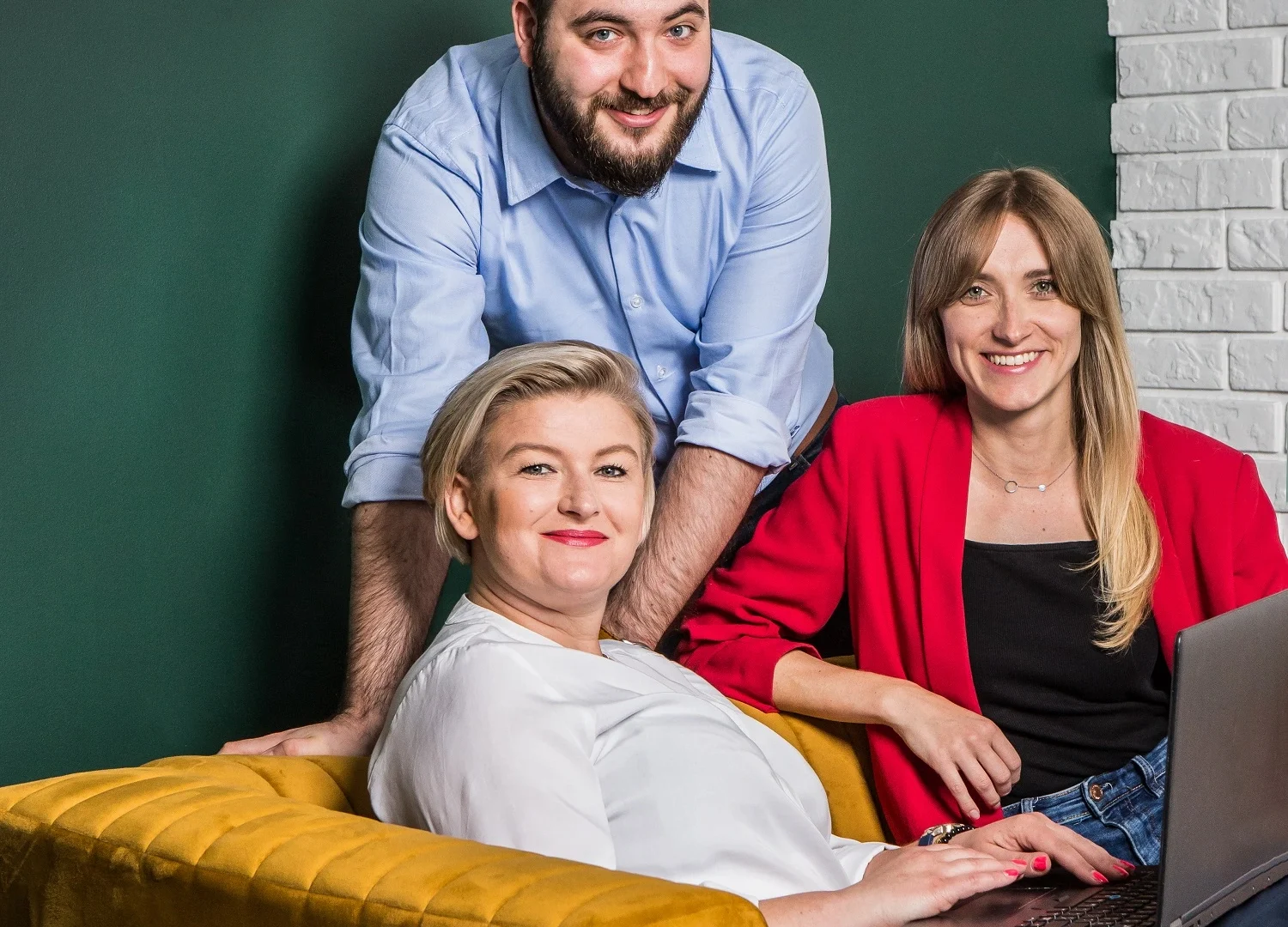 Zdjęcie biznesowe trzech uśmiechniętych osób w eleganckich, ale swobodnych stylizacjach, pracujących razem w kreatywnym biurowym otoczeniu. Kobieta w białej bluzce siedzi na musztardowej sofie, obok niej kobieta w czerwonej marynarce z laptopem, a za nimi stoi mężczyzna w niebieskiej koszuli, opierając się na oparciu. Zdjęcie symbolizuje współpracę, pozytywną atmosferę i profesjonalizm zespołu.